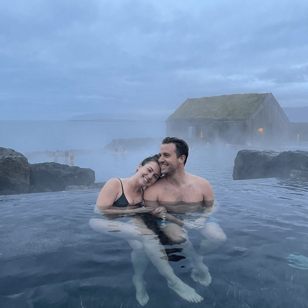 A couple smiling in a steaming geothermal lagoon with an ocean view.